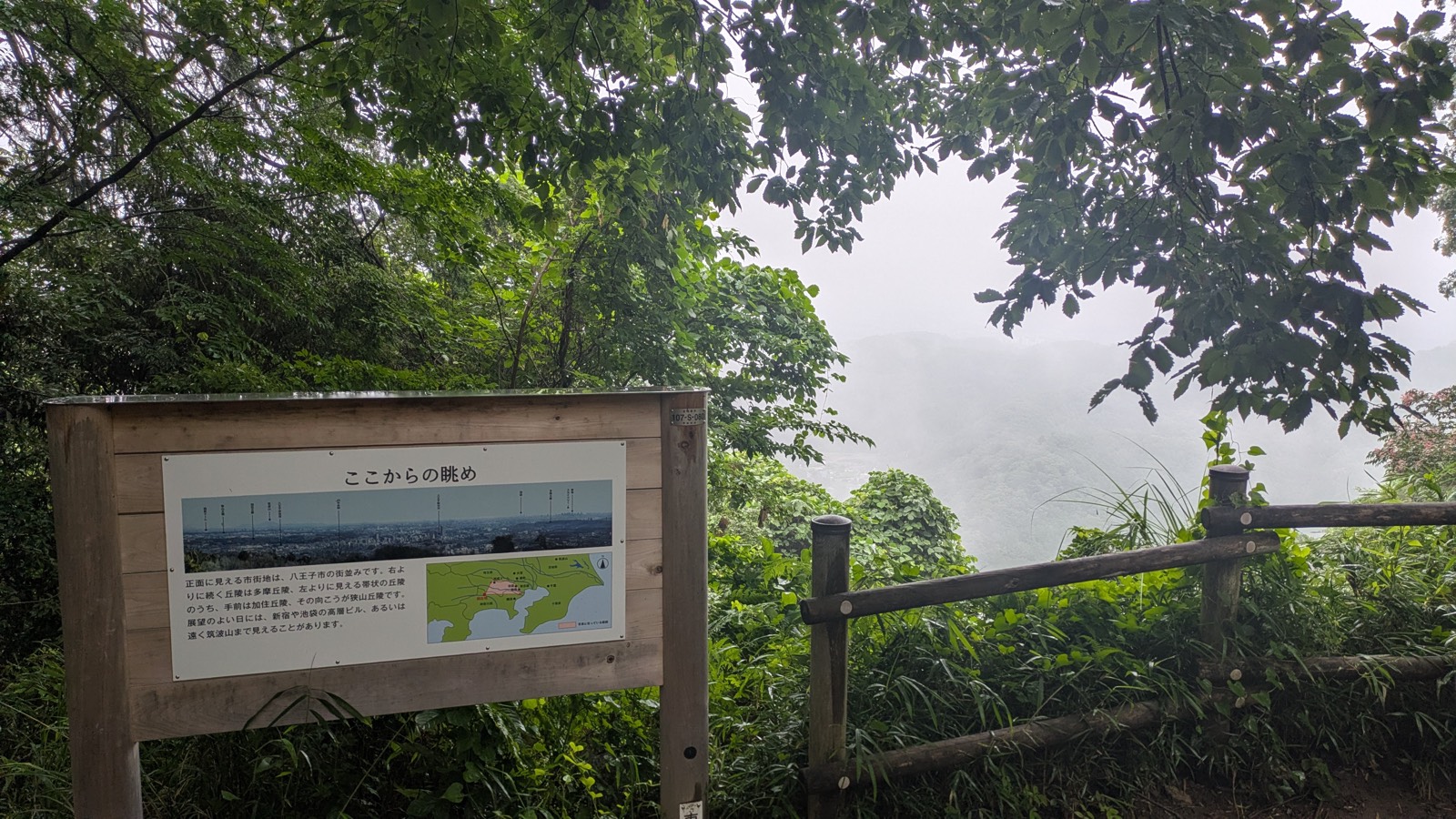 An interpretive board titled in Japanese at the Inariyama Observatory, with a labelled panorama photograph, beside a wooden fence in mist