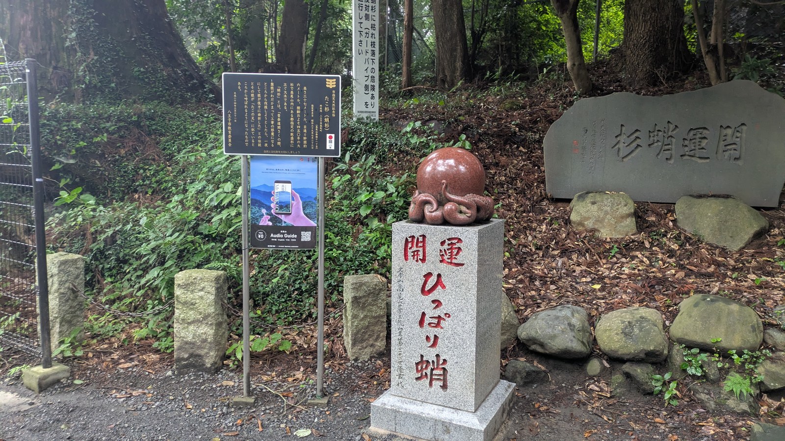 A polished red octopus statue on a granite plinth beside an information board on Mount Takao
