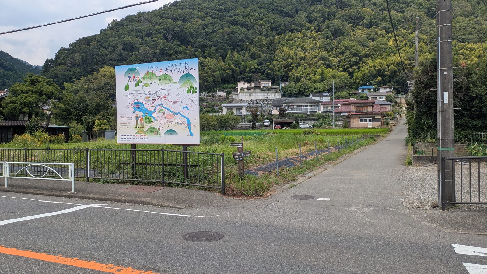 A village lane climbing away from a main road with a painted tourist map board beside it