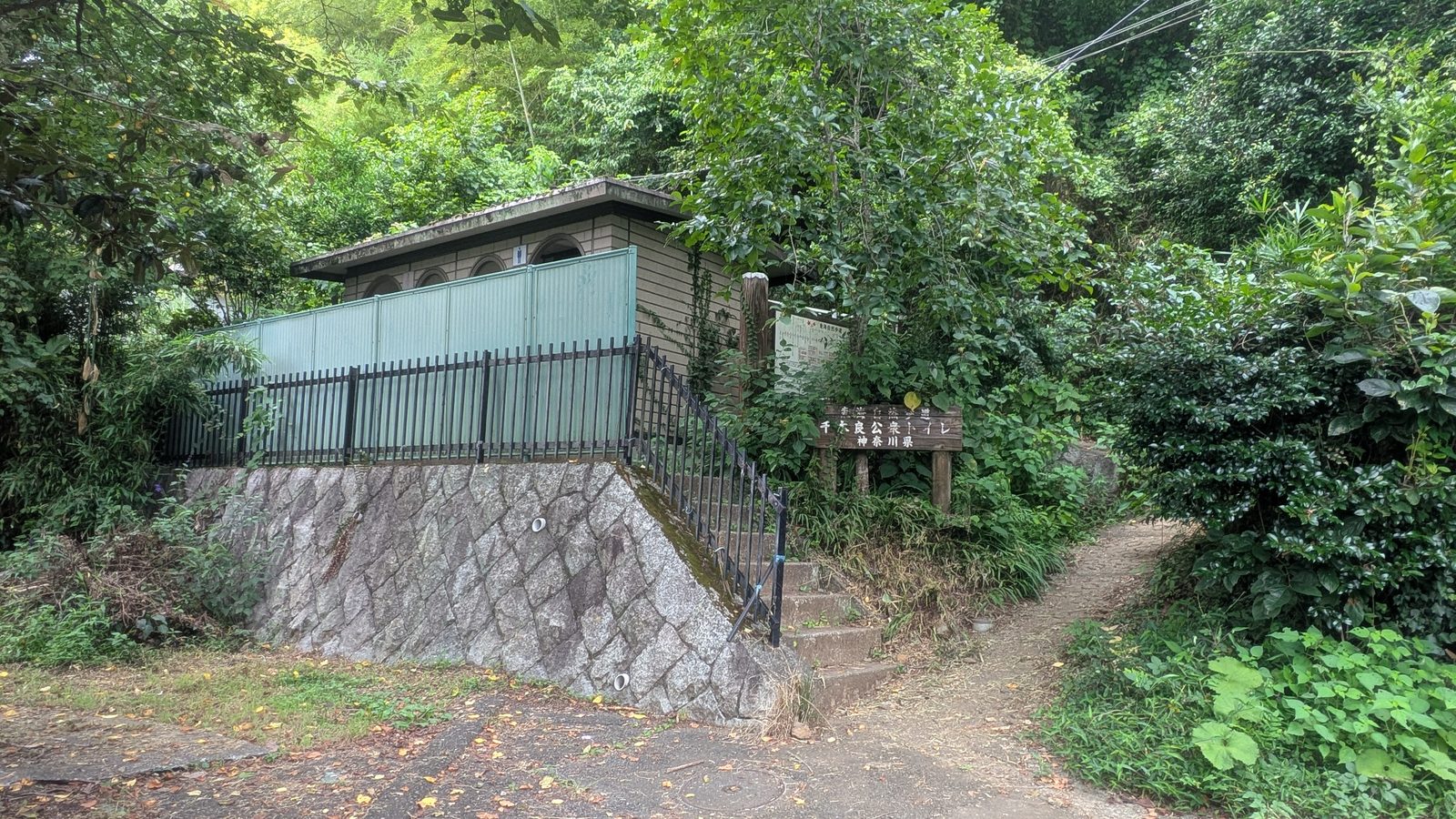 A small public toilet building beside a stone wall with a narrow path leading up into trees on its right