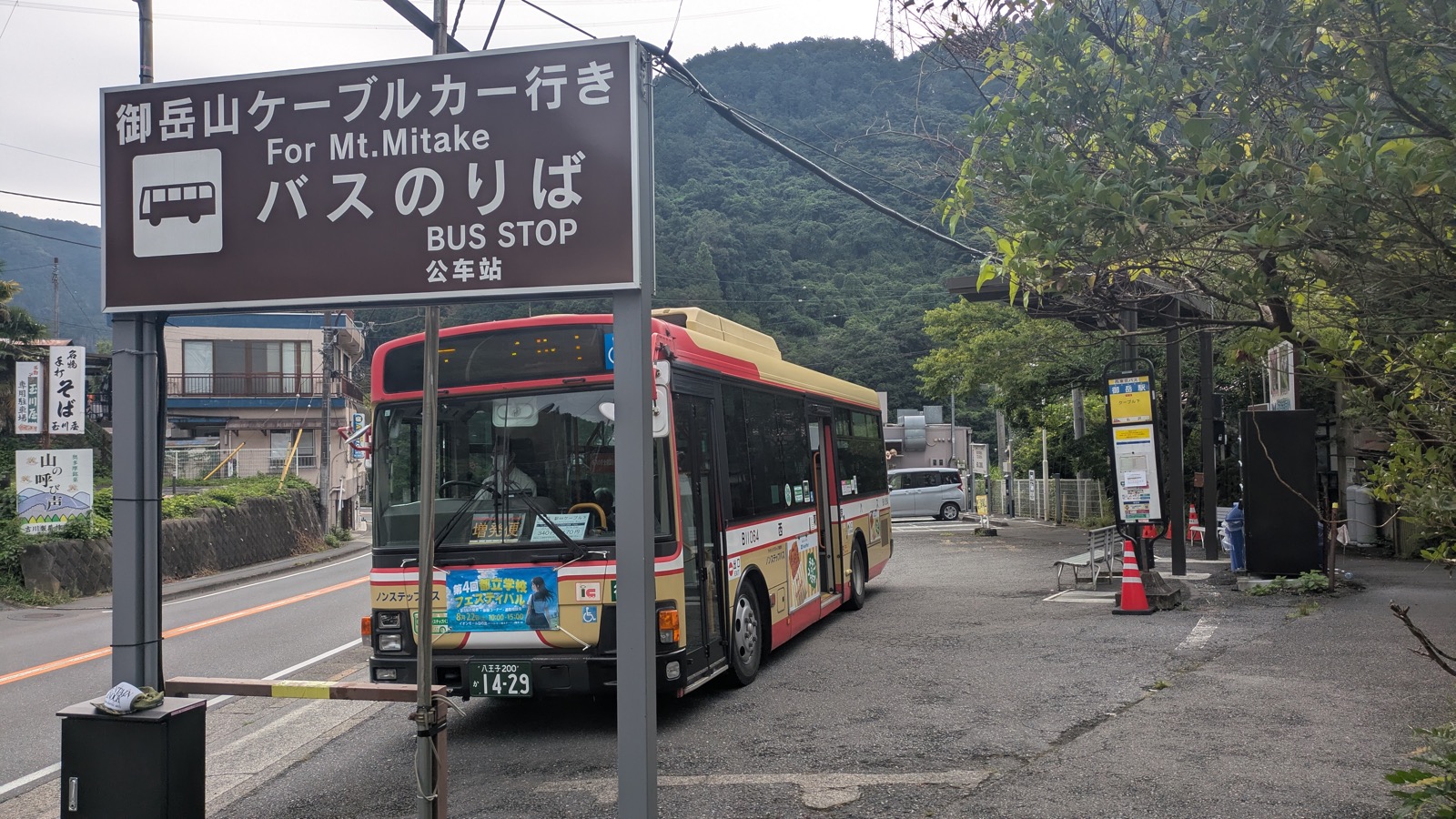 A red bus waiting at a rural stop beneath a brown sign reading For Mt Mitake BUS STOP
