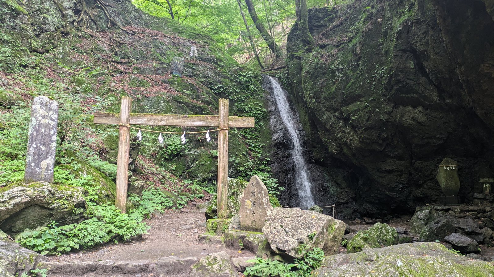 A wooden torii gate with a straw rope standing in front of a narrow waterfall in a rocky gorge