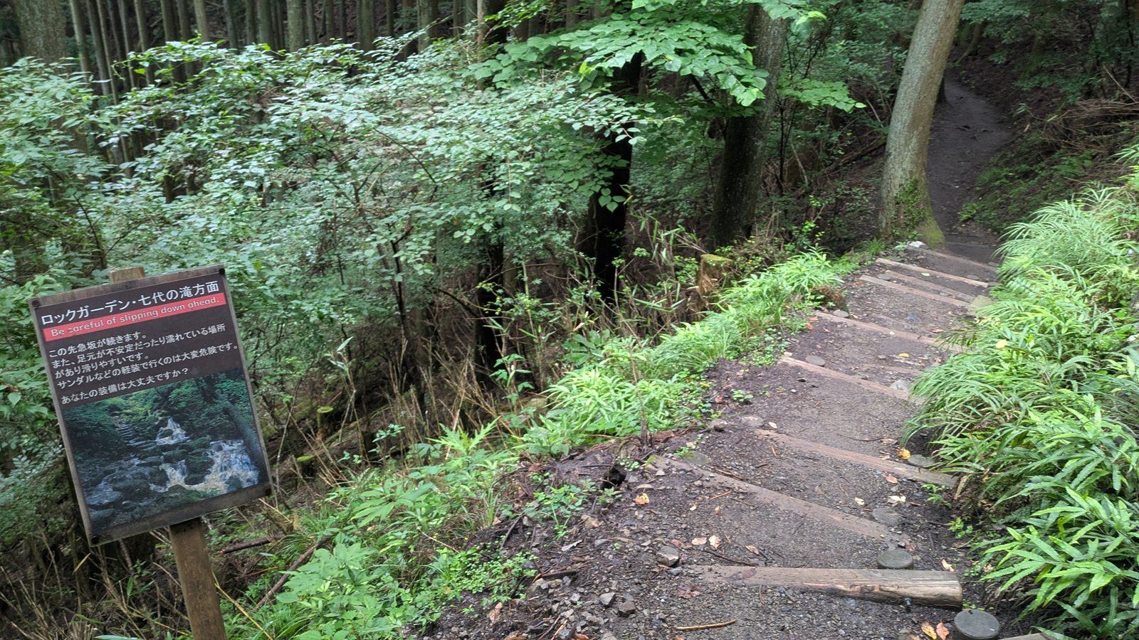 A brown warning sign with a red banner beside log steps descending into forest
