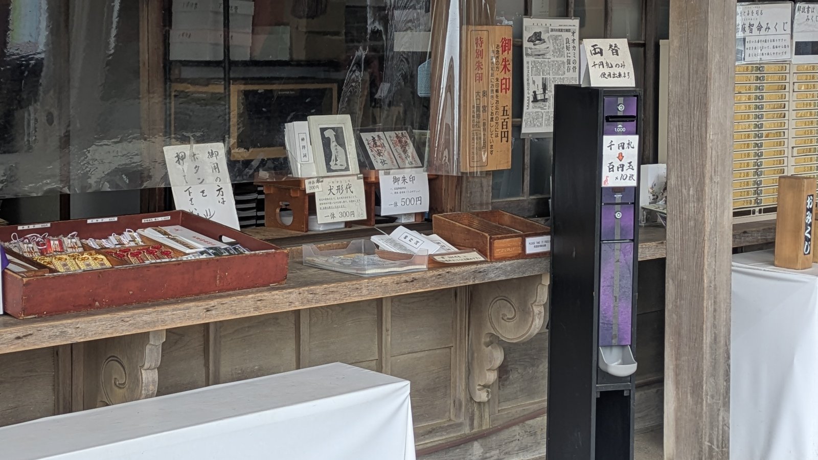 A shrine amulet counter with handwritten price cards beside a paper dog charm