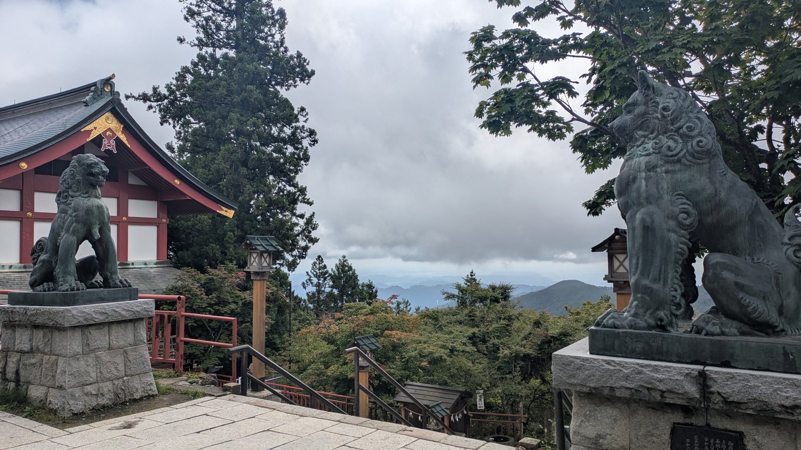 A view east over forested ridges from a mountain shrine, with a bronze guardian statue in the foreground