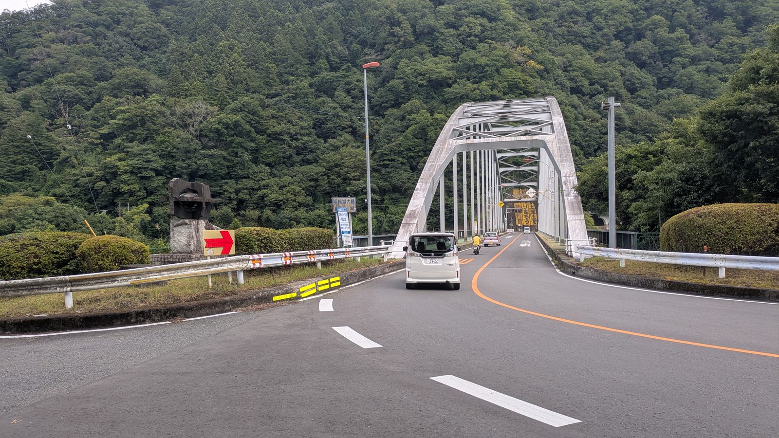 A steel arch road bridge crossing a river valley with wooded hills behind