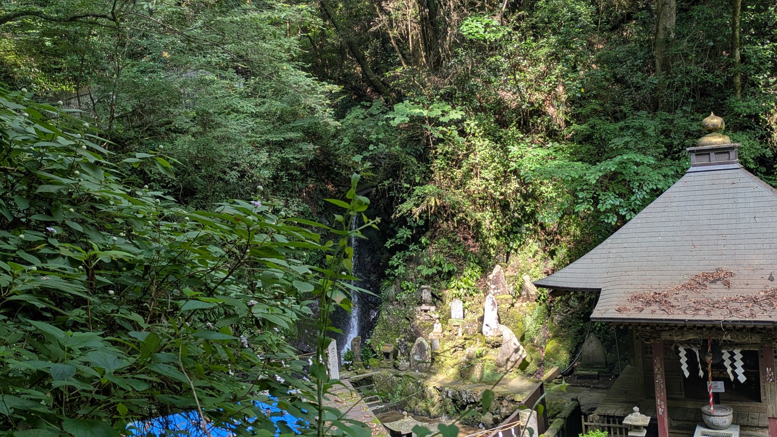 A view down from a forest trail onto a small temple hall with a narrow waterfall visible behind it