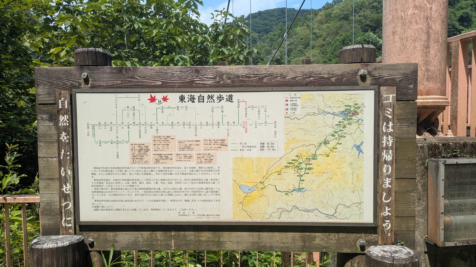 A large weathered wooden board displaying a long-distance trail route map beside a footbridge