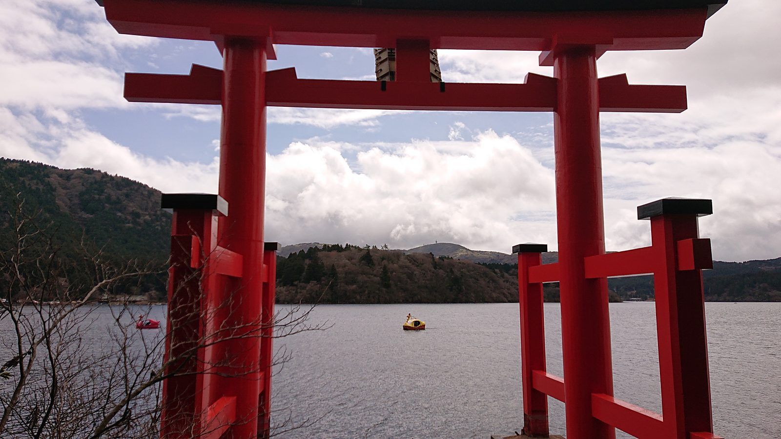 A red torii gate standing at the edge of Lake Ashi, with forested hills across the water
