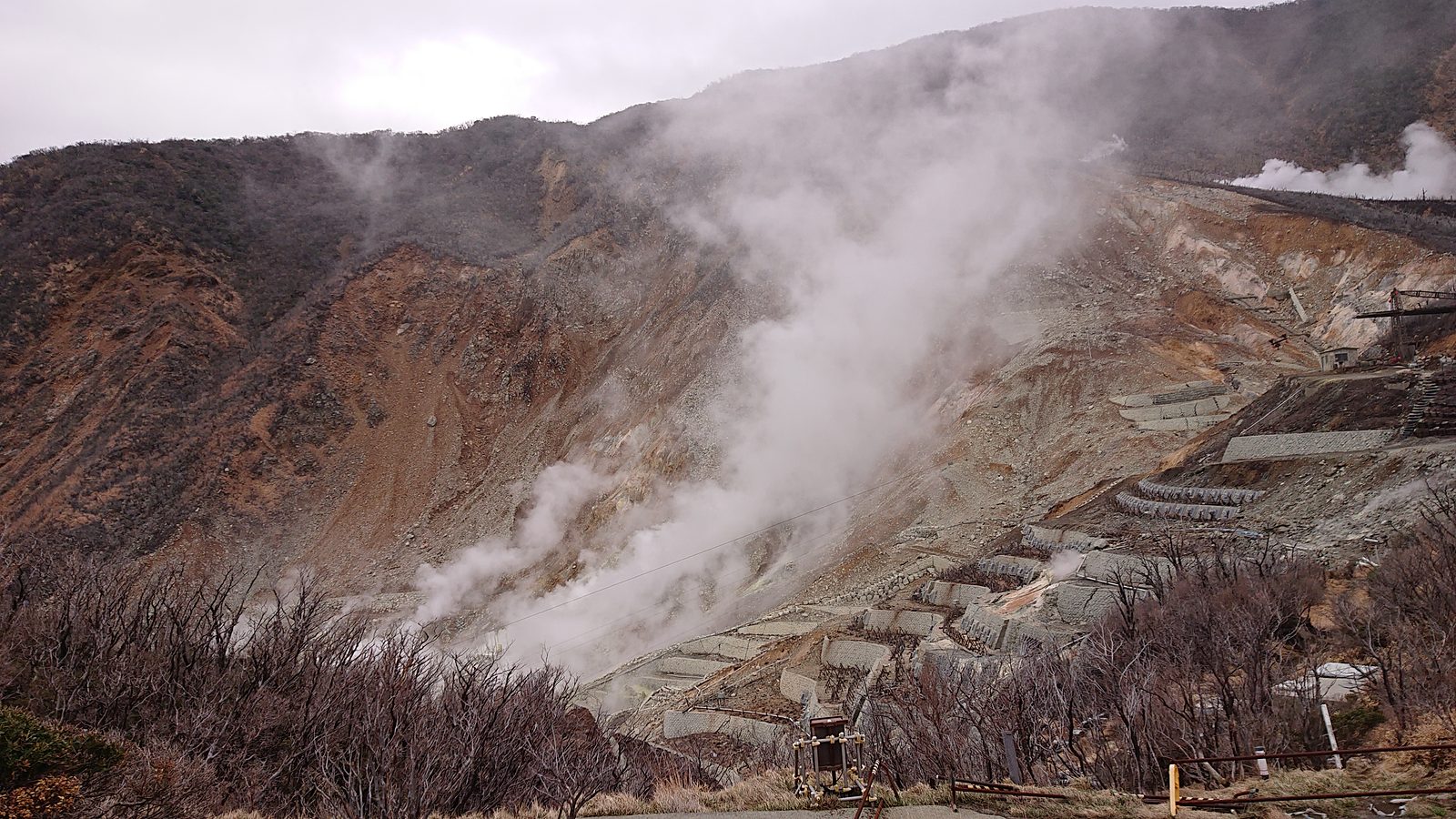 Steam rising from vents on a bare, rust-coloured mountainside at Owakudani, with rows of pipes on the slope
