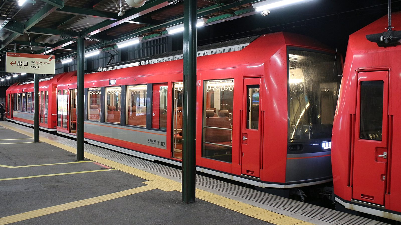 A red Hakone Tozan train at a covered platform, with a sign pointing to the cable car transfer, next to the exit