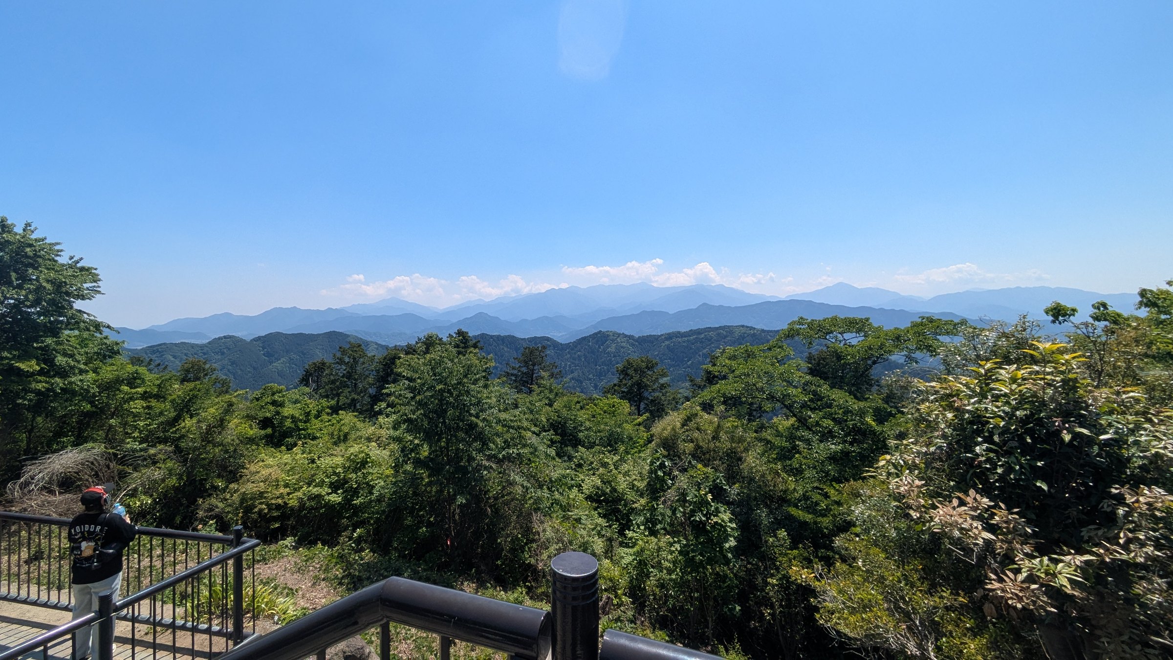 A walker at the rail of a viewing deck on the Minami-Takao ridge, looking out over green ridges to a line of hazy blue peaks with cumulus cloud building along the skyline