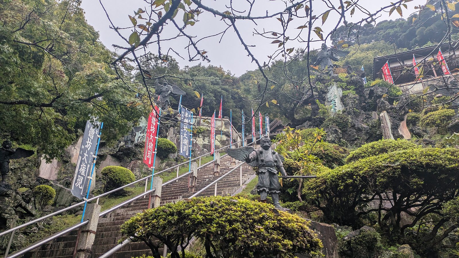 Stone steps lined with banners and a bronze crow tengu statue with wings on the slope