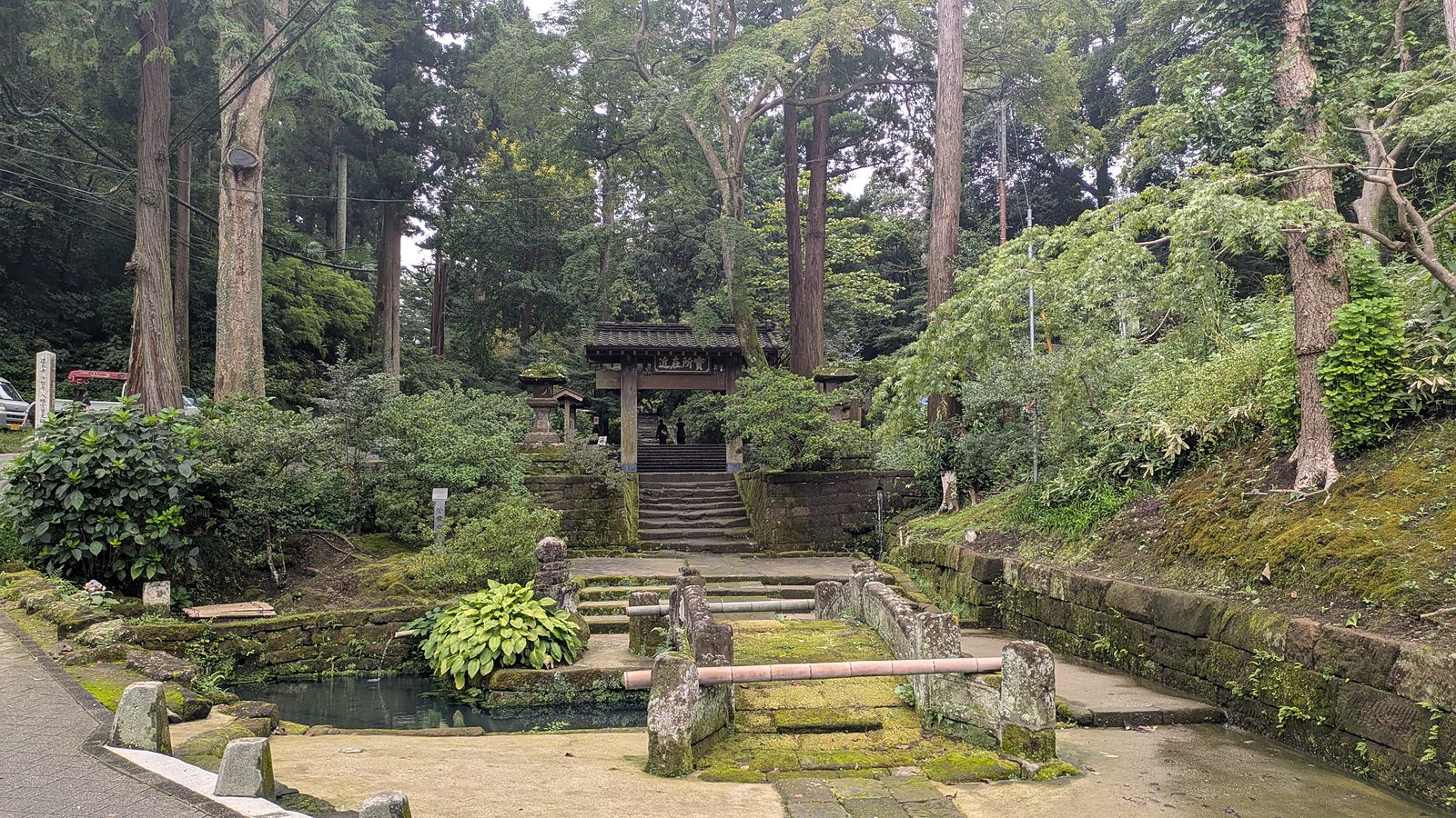 A stone bridge and a covered well in front of steps rising to a temple bell gate