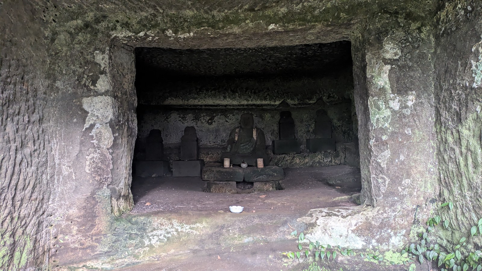 Inside a cave chamber, a seated stone figure with stone stupas on each side and offerings on a platform