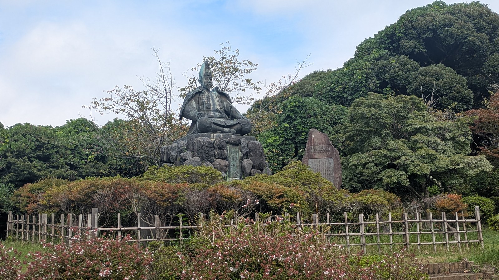 A seated bronze statue of Minamoto no Yoritomo on a rock base behind a bamboo fence
