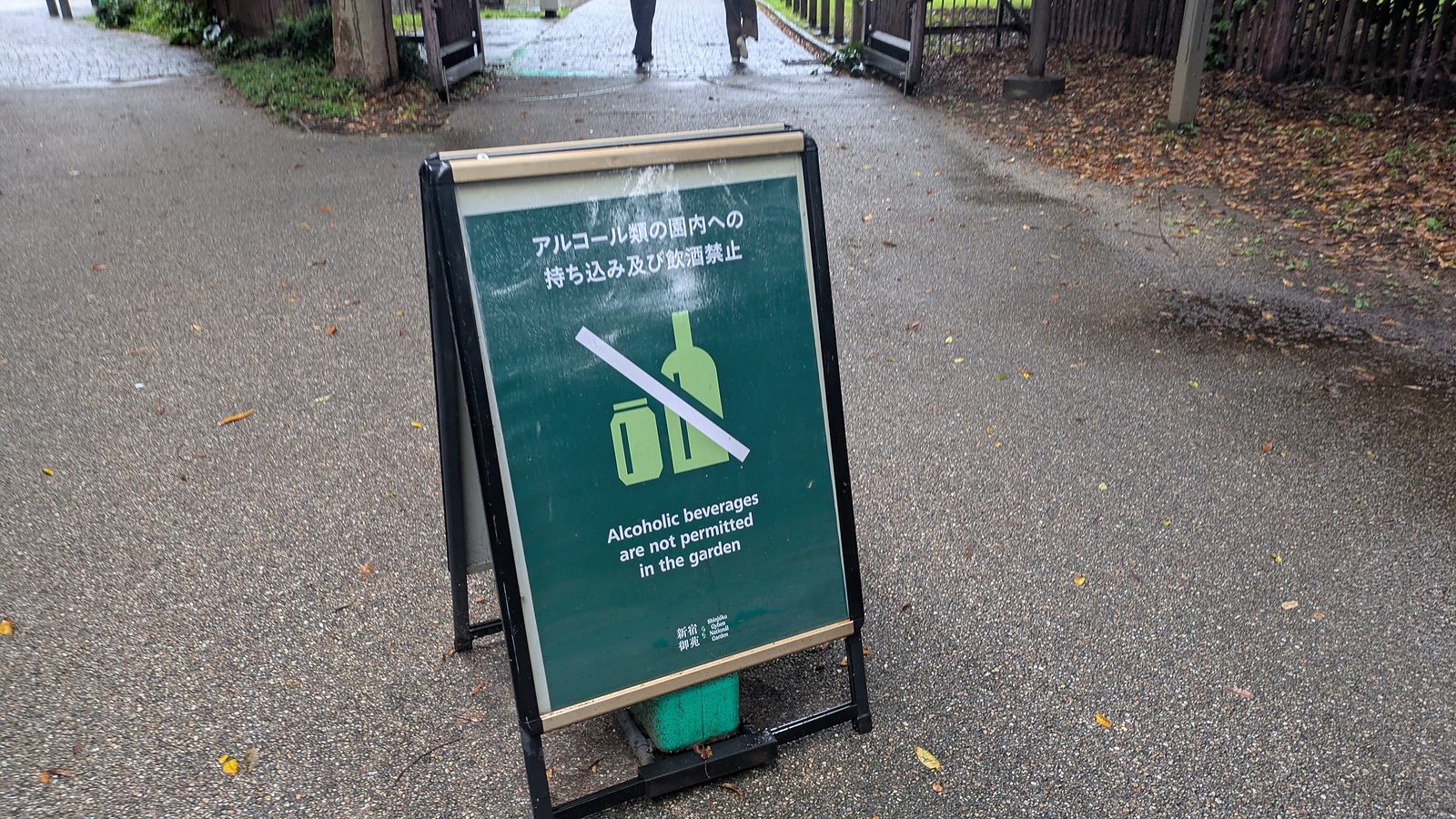 A green free-standing sign on a wet path showing a crossed-out bottle and the words Alcoholic beverages are not permitted in the garden