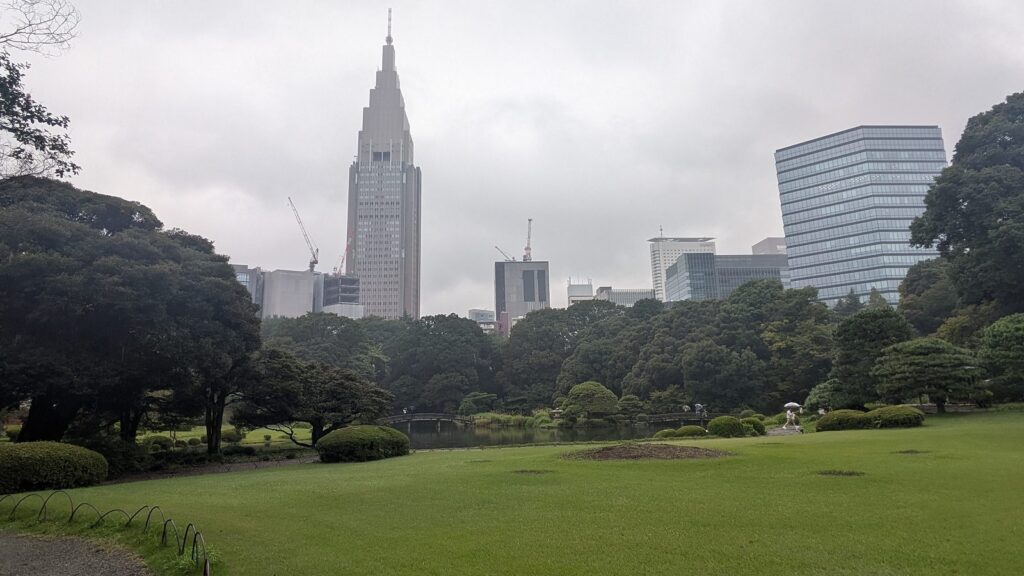 A pond with clipped pines in a Japanese garden, with the spire of a tall office tower rising behind the trees